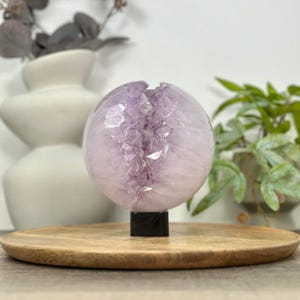 May include: A spherical amethyst crystal geode, displaying a mix of translucent and opaque purple hues. The crystal is mounted on a small black base, resting on a round wooden tray. Decorative items are in the background.