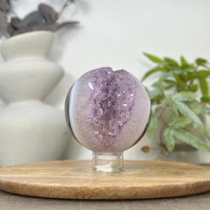 May include: A spherical amethyst geode crystal, showcasing a vibrant purple interior within a clear, polished exterior. The geode is mounted on a clear stand, set against a blurred background with decorative elements.