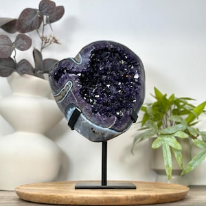May include: A deep purple amethyst geode with jagged, crystalline interior structures and a smooth, greyish-brown outer rock surface. The geode is securely mounted on a thin, black metal stand with a square base, positioned upright for display.