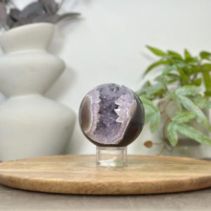 May include: A polished, spherical amethyst geode, showcasing a vibrant purple crystal interior. The geode is set on a clear stand, resting on a round wooden tray. Decorative elements include a white vase and potted greenery.