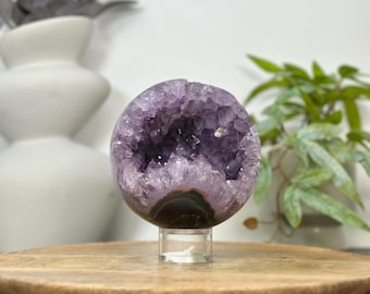 May include: A spherical amethyst geode, showcasing a deep purple interior with crystal formations. The geode is mounted on a clear stand, set on a wooden tray. The geode is a natural home decor item.