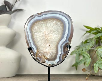 May include: A polished agate slice featuring concentric bands of light blue, gray, and white surrounding a central crystalline geode cavity. The stone is mounted on a black metal stand with a square base, displaying a natural mineral formation.