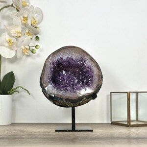 May include: A circular amethyst geode featuring deep purple crystalline structures within a natural rock formation, mounted on a black metal stand. The geode displays a polished outer edge and a rough, textured exterior, showcasing natural mineral variations.