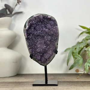 May include: A vertical amethyst geode slice with deep purple crystalline structures and a dark outer rim, mounted on a black metal stand with a square base. The crystal surface features numerous small, reflective points of light.