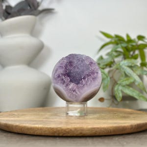 May include: A spherical amethyst geode, showcasing a vibrant purple interior and a banded, light-colored exterior. The geode is displayed on a clear acrylic stand, set against a wooden surface, with a blurred background of decorative vases and greenery.