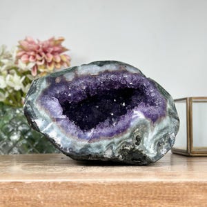 May include: A large amethyst geode with a deep purple crystal interior. The outer shell is a mix of gray, green, and white. The geode sits on a wooden surface, with flowers and a decorative box in the background.