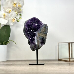 May include: A large amethyst geode crystal on a black metal stand. The geode features a deep purple crystalline interior and a gray and blue exterior. The stand is square and the geode is displayed in front of a white background.