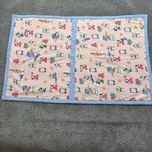 Puede incluir: Una manta acolchada rectangular con un fondo de rayas rosas y blancas, decorada con un patrón repetitivo de Mickey Mouse, globos y estrellas. La manta está bordeada por una cenefa de tela azul claro e incluye cintas de color azul.