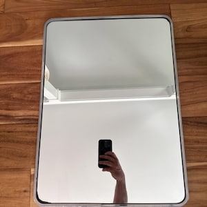 Puede incluir: Un espejo de pared rectangular con esquinas redondeadas y un marco fino de metal plateado. La superficie reflectante muestra una vista clara de un techo blanco, una moldura de pared blanca y un teléfono inteligente negro en una mano.