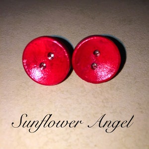 Op de afbeelding: Een paar rode houten oorbellen met twee kleine zilveren studs op elke oorbel. De oorbellen zijn rond en hebben een glanzende afwerking.