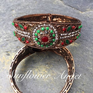 May include: A copper bangle bracelet with a decorative design featuring red, green, and white stones. The bracelet has a hinged clasp.