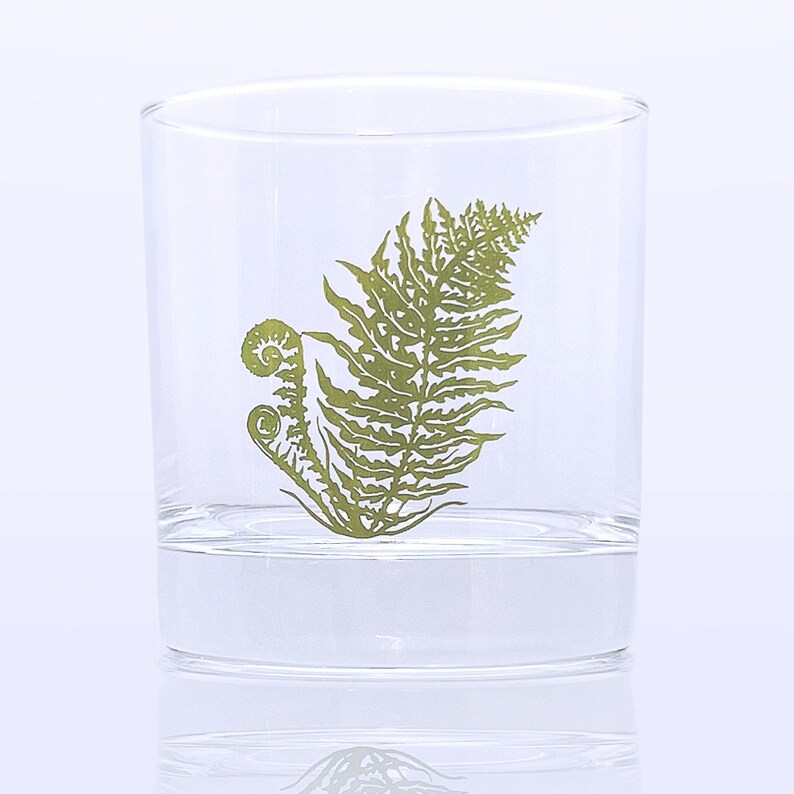 K&ouml;nnte beinhalten: Ein durchsichtiger Glasbecher mit einem gr&uuml;nen Farnmuster. Der Farn ist stilisiert und hat eine eingerollte Wedel.