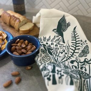 May include: A white tea towel with a green print of a bird, ferns, mushrooms, and other forest elements.