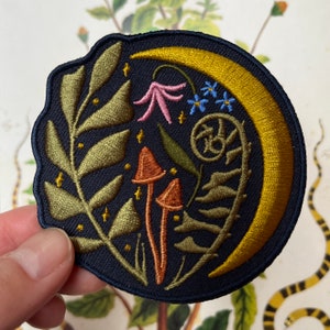 May include: Embroidered patch featuring a crescent moon, mushrooms, leaves, and flowers on a dark blue background. The moon is gold, the mushrooms are orange and brown, the leaves are green, and the flowers are pink and blue.