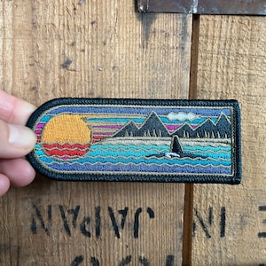 May include: A colourful embroidered patch depicting a sunset scene over a body of water. The patch features a large orange sun, mountains, and a whale swimming in the water. The patch is surrounded by a dark blue border.