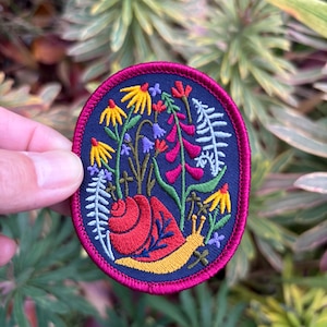 Peut inclure: Un patch brodé coloré représentant un escargot entouré de fleurs et de feuilles. L'escargot est rouge et jaune, et les fleurs sont jaunes, rouges, bleues et violettes. Le patch est circulaire et a un fond bleu foncé avec une bordure rose.