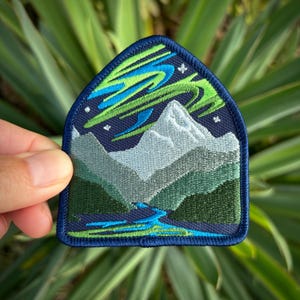 May include: Embroidered patch depicting a mountain range with a river flowing through it, under a dark blue sky with green and blue aurora borealis. The patch has a navy blue border.