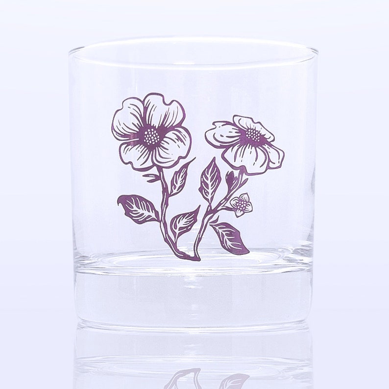 K&ouml;nnte beinhalten: Ein klares Glas mit einem violetten Blumenmuster. Die Blumen sind wei&szlig; mit violetten Umrissen und Stielen. Das Glas ist perfekt zum Servieren von Getr&auml;nken.