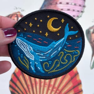 May include: A circular embroidered patch depicting a blue whale in the sea beneath a night sky with a yellow crescent moon and stars. The design includes stylized waves and has a black border.