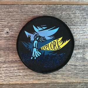 May include: A black and blue embroidered patch featuring a girl holding a torch and a banner that reads "EXPLORE".