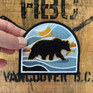 May include: An embroidered patch depicting a black bear with a fish in its mouth, standing in blue water with a yellow sun in the background.