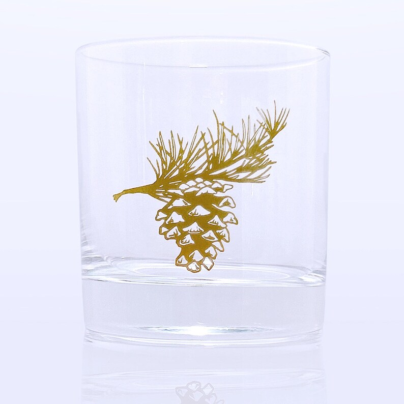 K&ouml;nnte beinhalten: Ein klares Glas mit einem goldenen Kiefernzapfen- und Zweigdesign.