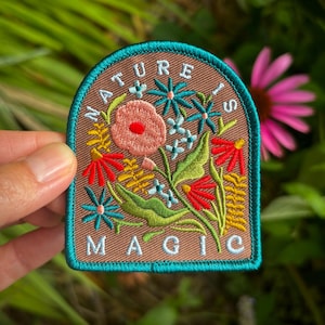 May include: A brown patch with a teal border featuring a floral design and the words "NATURE IS MAGIC". The patch includes a pink poppy, red and blue flowers, and green leaves. The patch is held in front of a blurred green background.