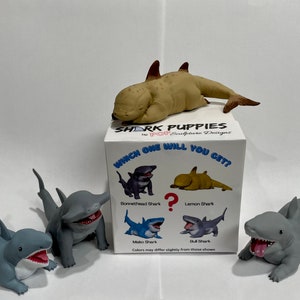 May include: A set of five shark puppy figurines in various poses, including a bonnethead shark, a mako shark, a lemon shark, a bull shark, and a grey shark. The figurines are all in a playful, cartoonish style and are made of a soft, rubbery material. The figurines are packaged in a white box with the words "Shark Puppies" and "Which one will you get?" printed on it.