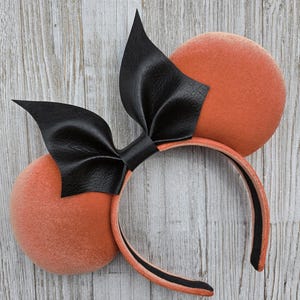 May include: Orange mouse ear headband with a black leather bow. The ears are round and plush, and the headband is black and orange. The bow is a prominent feature, adding a stylish touch to the accessory.