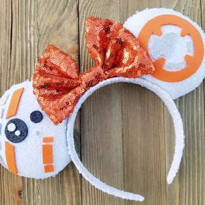 May include: White sequined headband with BB-8 inspired ears. The ears feature orange and silver accents, with a large orange sequined bow. The design is inspired by the Star Wars character BB-8.