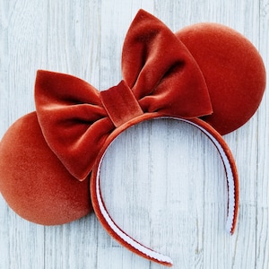 May include: A pair of burnt orange velvet Minnie Mouse ears with a white headband. The ears are large and floppy, and the headband is adjustable.