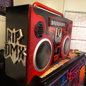 Puede incluir: Un gran boombox rojo y negro con un logotipo plateado "RIP DMX" en el lateral. El boombox tiene un altavoz grande a cada lado y un altavoz más pequeño en la parte superior. El boombox está sentado en una mesa con otro boombox en el fondo.