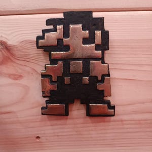 Peut inclure: Une figurine pixelisée noire et dorée, ressemblant à un personnage de jeu vidéo, est montée sur une surface en bois.