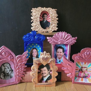 May include: A collection of six small, ornate picture frames with various images of people. The frames are made of gold, pink, and blue metal and have a variety of designs, including hearts, stars, and flowers. The images in the frames include a man in a suit, a woman in a dress, a man with a beard, a woman with blonde hair, and a woman with brown hair.
