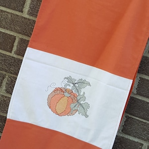 以下が含まれることがあります： 刺繍されたカボチャのデザインが施された白いポケットが付いたオレンジ色のエプロン。エプロンは首周りに黄色のトリムがあり、レンガの壁を背景に木製のハンガーに吊るされています。