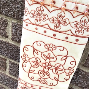 May include: Close-up of a cream fabric sleeve with detailed rust-coloured embroidery. The design includes floral patterns and beaded accents along the top edge. The sleeve is set against a brick wall.