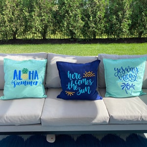 K&ouml;nnte beinhalten: Drei dekorative Zierkissen auf einem grauen Outdoor-Sofa. Die Kissen sind t&uuml;rkis und marineblau, mit Text und Grafiken zum Thema Tropen. Ein Kissen sagt "Aloha Summer" mit einer Ananas, ein anderes sagt "Here comes the sun", und das dritte sagt "Yes way, Vacay!"