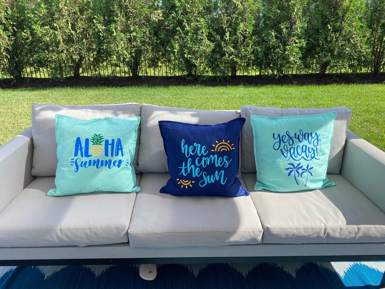 K&ouml;nnte beinhalten: Drei dekorative Zierkissen auf einem grauen Outdoor-Sofa. Die Kissen sind t&uuml;rkis und marineblau, mit Text und Grafiken zum Thema Tropen. Ein Kissen sagt "Aloha Summer" mit einer Ananas, ein anderes sagt "Here comes the sun", und das dritte sagt "Yes way, Vacay!"