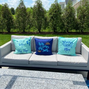 K&ouml;nnte beinhalten: Ein graues Outdoor-Sofa mit drei dekorativen Kissen. Die Kissen sind hellblau und marineblau, mit Texten wie "Life is better in Flip Flops", "Here comes the sun" und "Yes way vacay". Das Sofa steht auf einer Terrasse.