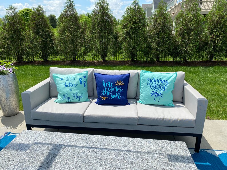 K&ouml;nnte beinhalten: Ein graues Outdoor-Sofa mit drei dekorativen Kissen. Die Kissen sind hellblau und marineblau, mit Texten wie "Life is better in Flip Flops", "Here comes the sun" und "Yes way vacay". Das Sofa steht auf einer Terrasse.