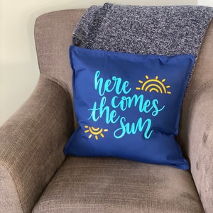 K&ouml;nnte beinhalten: Ein marineblaues Zierkissen mit dem Schriftzug "here comes the sun" in hellblauer Schrift und einer gelben Sonnengrafik. Das Kissen liegt auf einem braunen Sessel mit einer grauen Decke.
