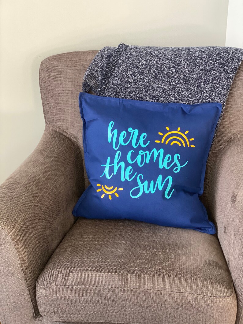 K&ouml;nnte beinhalten: Ein marineblaues Zierkissen mit dem Schriftzug "here comes the sun" in hellblauer Schrift und einer gelben Sonnengrafik. Das Kissen liegt auf einem braunen Sessel mit einer grauen Decke.