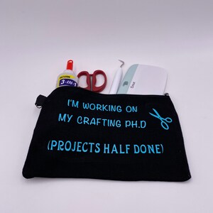 Op de afbeelding: Zwarte ritszak met de tekst "Ik werk aan mijn handwerk-doctoraat (Projecten half klaar)" in blauwe letters. Naast de tekst is een schaar afgebeeld. De tas is gevuld met knutselspullen, waaronder een rode schaar, een witte schraper, een wit onkruidverwijderingsgereedschap en een fles lijm.