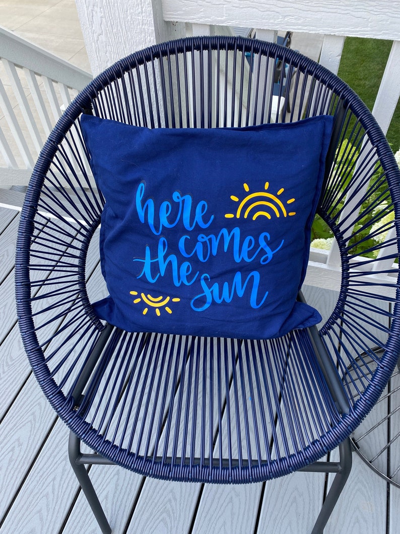 K&ouml;nnte beinhalten: Ein marineblaues Kissen mit dem Text "here comes the sun" in blauer Schrift und gelben Sonnengrafiken. Das Kissen liegt auf einem marineblauen Webstuhl mit Metallrahmen, auf einer grauen Terrasse.
