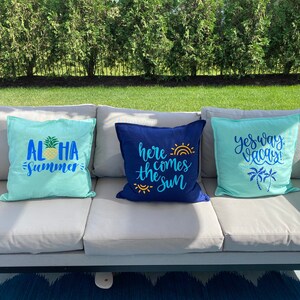 K&ouml;nnte beinhalten: Drei dekorative Wurfkissen auf einem grauen Outdoor-Sofa. Ein hellblaues Kissen hat die Worte "ALOHA SUMMER" mit einer Ananas-Grafik. Ein marineblaues Kissen sagt "here comes the sun". Ein weiteres hellblaues Kissen sagt "yes way vacay!"