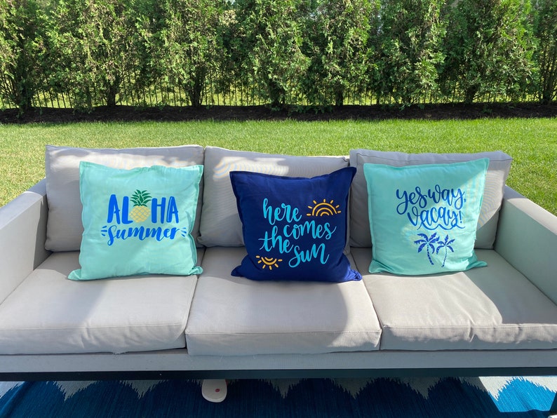K&ouml;nnte beinhalten: Drei dekorative Wurfkissen auf einem grauen Outdoor-Sofa. Ein hellblaues Kissen hat die Worte "ALOHA SUMMER" mit einer Ananas-Grafik. Ein marineblaues Kissen sagt "here comes the sun". Ein weiteres hellblaues Kissen sagt "yes way vacay!"