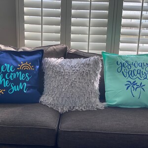 K&ouml;nnte beinhalten: Drei dekorative Kissen auf einem grauen Sofa. Ein marineblaues Kissen hat den Text "here comes the sun" in T&uuml;rkis und Gelb. Ein flauschiges, graues Kissen befindet sich in der Mitte. Ein hellblaues Kissen hat den Text "yes way, vacay!" mit Palmen.