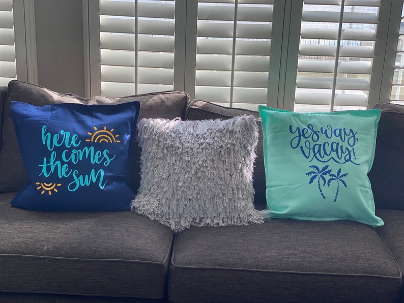 K&ouml;nnte beinhalten: Drei dekorative Kissen auf einem grauen Sofa. Ein marineblaues Kissen hat den Text "here comes the sun" in T&uuml;rkis und Gelb. Ein flauschiges, graues Kissen befindet sich in der Mitte. Ein hellblaues Kissen hat den Text "yes way, vacay!" mit Palmen.