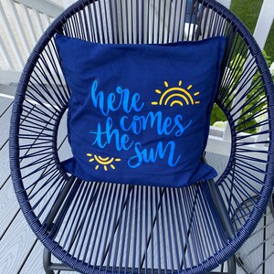 K&ouml;nnte beinhalten: Ein marineblaues Zierkissen mit dem Schriftzug "here comes the sun" in blauer Schrift und einer gelben Sonne. Das Kissen liegt auf einem marineblauen Webstuhl. Der Stuhl steht auf einer Holzterrasse, was ein harmonisches Bild ergibt.
