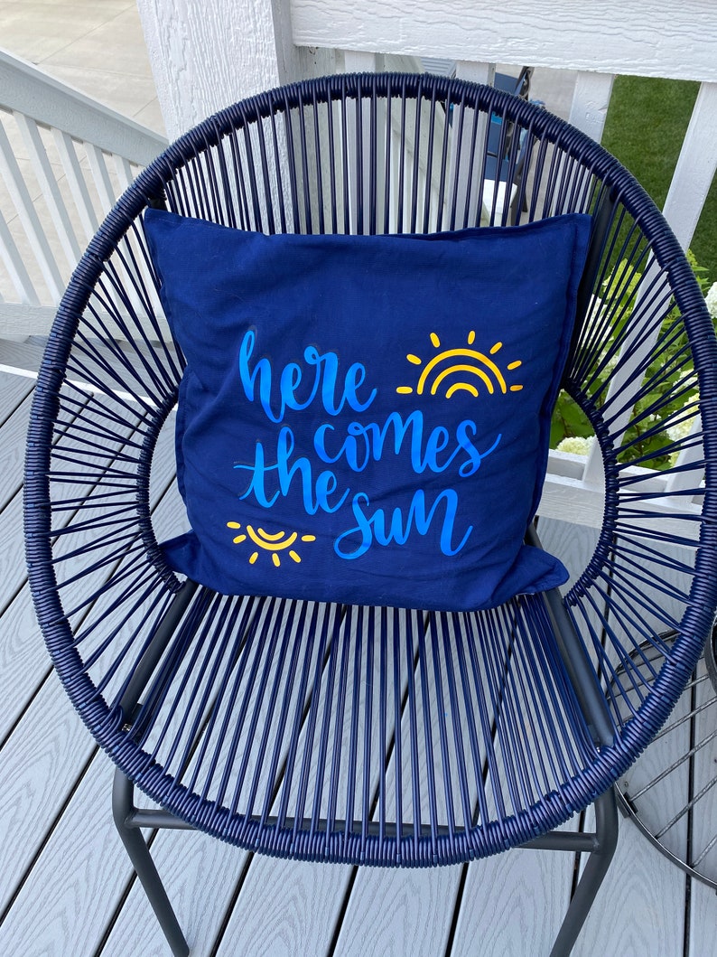 K&ouml;nnte beinhalten: Ein marineblaues Zierkissen mit dem Schriftzug "here comes the sun" in blauer Schrift und einer gelben Sonne. Das Kissen liegt auf einem marineblauen Webstuhl. Der Stuhl steht auf einer Holzterrasse, was ein harmonisches Bild ergibt.